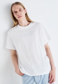 Balta medvilnės marškinėliai su iškilniu "LEVI'S" logotipu priekyje, apvaliu kaklu, laisvu siluetu, dėvimi su šviesiai mėlynomis džinsinėmis šortais.