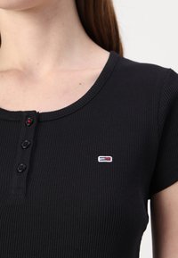 Fekete bordázott rövidujjú felső kerek nyakkivágással, három gombbal és egy kis logóval a mellkasán.