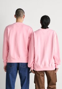 Dos personas llevan sudaderas rosas de gran tamaño. Las sudaderas tienen puños de canalé y un cuello redondeado. El fondo es neutro.