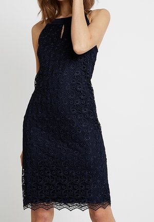 Granatowa sukienka na ramiączkach z koronki z okrągłymi i kwiatowymi wzorami, małym wycięciem z przodu i falbankowanym dołem, noszona przez stojącą kobietę.