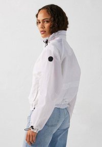 Witte windbreakerjas met een hoge kraag, koordsluitingen bij de mouwen en een zwart logopatch op de linkermouw. Lichtgewicht, gestructureerde stof.
