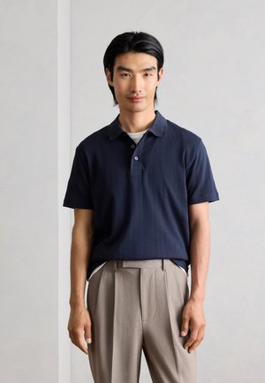 Junger Mann mit schwarzen Haaren, der ein marineblaues kurzärmeliges Poloshirt und beige Hosen trägt, steht vor einem hellgrauen Hintergrund.