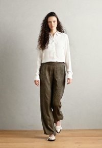 Witte overhemd met lange mouwen, groene patroonbroek en witte instap schoenen met een gesp. Model staat tegen een neutrale achtergrond.