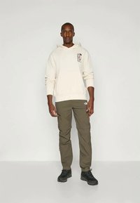 Άνδρας που φοράει λευκή μπλούζα, ελαιόχρωμα cargo παντελόνια και μαύρες ορειβατικές μπότες, στέκεται μπροστά σε απλό ανοιχτό φόντο.