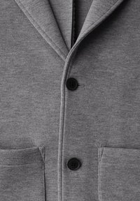 Harmaa neuletakki, jossa on läppäkaulus, kaksi etutaskua ja yksi musta nappisulku. Valmistettu pehmeästä, teksturoidusta kankaasta.