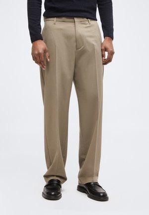 Mann trägt beige weit geschnittene Hosen, schwarze Leder-Loafer und ein schwarzes langärmeliges Oberteil, steht vor einem weißen Hintergrund.