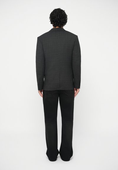 Mann mit lockigem Haar, der ein dunkelgraues karierter Blazer und schwarze weiter geschnittene Hose trägt, und vor einem weißen Hintergrund steht.