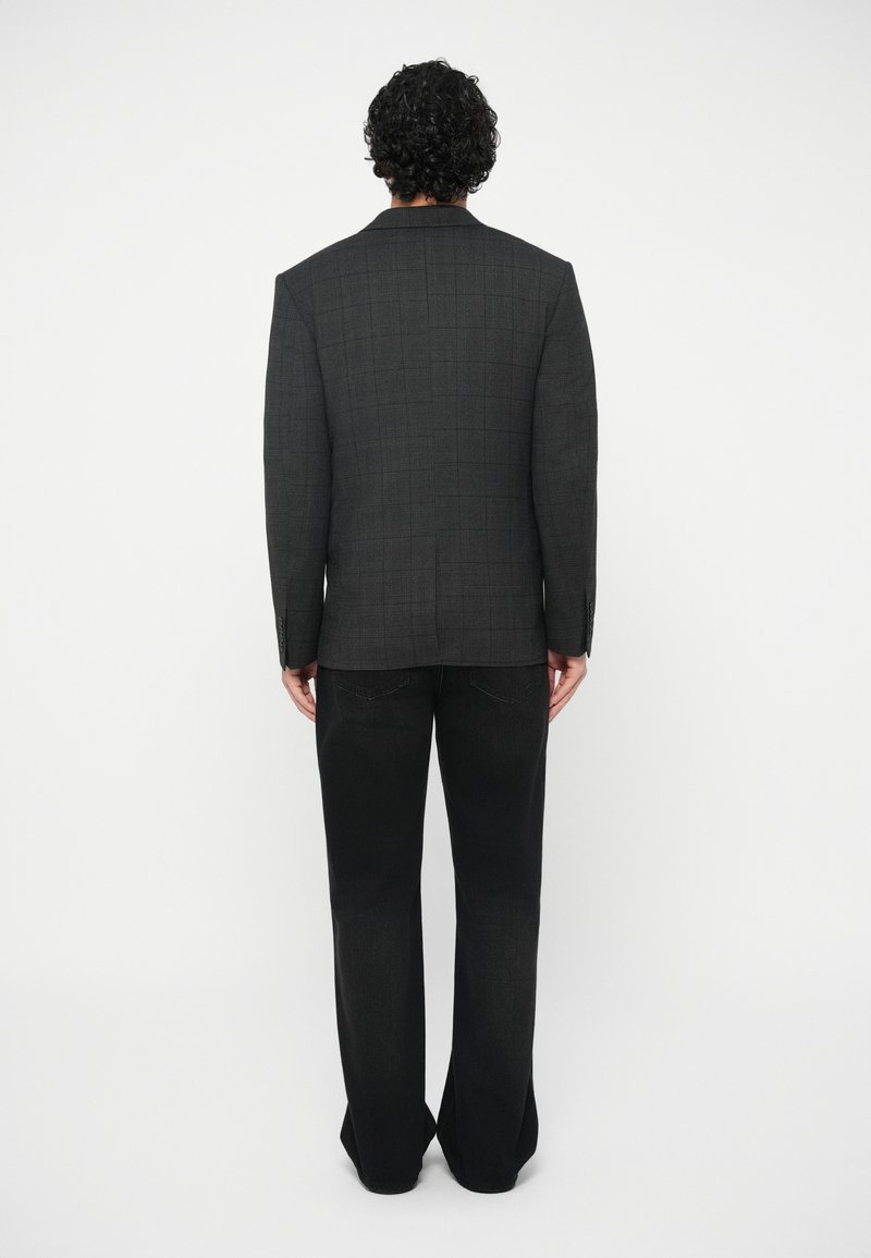 Mann mit lockigem Haar, der ein dunkelgraues karierter Blazer und schwarze weiter geschnittene Hose trägt, und vor einem weißen Hintergrund steht.