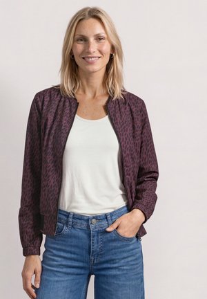 Femme blonde portant une veste bordeaux à motifs avec zip sur une chemise blanche et un jean bleu, souriante, une main dans la poche.