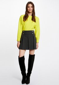 Maglione giallo lavorato a maglia con maniche lunghe abbinato a una gonna a pieghe grigia scura a vita alta con dettagli in bottoni argentati e stivali neri alti fino alla coscia.