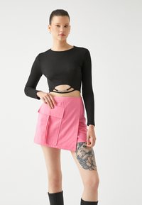 Musta pitkähihainen crop-top, jossa on ribbikuvio ja aukko-yksityiskohta; vaaleanpunainen mini-hame etutaskulla ja sivuhalkiolla; malli, joka käyttää polven yli ulottuvia saappaita.