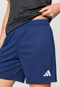 Μπλε αθλητικό σορτς με ελαστική μέση, κατασκευασμένο από λείο ύφασμα. Διαθέτει λε υκό λογότυπο Adidas στο κάτω αριστερό μέρος.