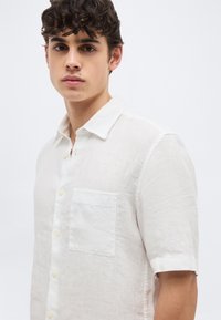 Νέος άνδρας με σκούρα σγουρά μαλλιά φοράει λευκό πουκάμισο με κοντά μανίκια και τσέπη στο στήθος, στέκεται μπροστά σε απλό ανοιχτό φόντο.