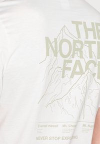 Bílý tričko s grafikou světle zelených hor a textem na zadní straně, zobrazující různé názvy hor a jejich souřadnice.