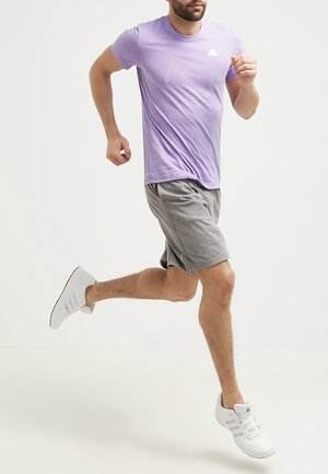 Camiseta atlética de manga corta en color púrpura con tejido texturizado, combinada con pantalones cortos grises. El modelo lleva zapatillas blancas con tres franjas.