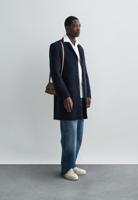 Navy wool coat with buttons, white collared shirt, light blue jeans, and cream sneakers. Brown shoulder bag adds contrast.