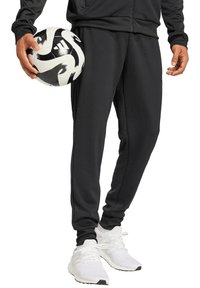 Zwarte sportieve sweatpants met een taps toelopend ontwerp en zijzakken, gecombineerd met witte gestructureerde sneakers. Een zwart-wit voetbal in de hand gehouden.