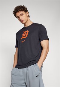 Tamsiai pilkas trumpomis rankovėmis marškinėliai su oranžiniu „D“ logotipu ir mažu Nike ženkliuku. Dėvimi su šviesiai pilkais šortais. Lygus audinio tekstūras.
