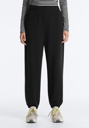 Person trägt schwarze Jogginghosen mit elastischem Bund, ein gestreiftes Langarmshirt und beige Sneaker mit neongelben Schnürsenkeln und steht vor weißem Hintergrund.