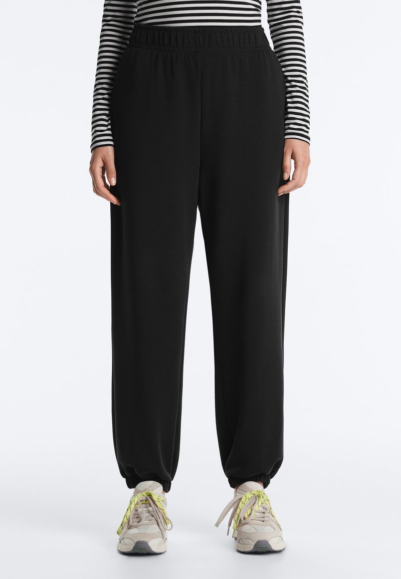 Osoba nosiaca čierne tepláky s elastickým pásom, pruhované tričko s dlhým rukávom a béžové tenisky s neónovo žltými šnúrkami, stojaca na bielom pozadí.