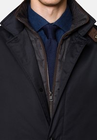 Czarna kurtka zewnętrzna z brązowymi akcentami na kołnierzu, noszona na ciemnoniebieskiej koszuli i teksturowanym granatowym krawacie, z widocznym detalem zamka błyskawicznego.