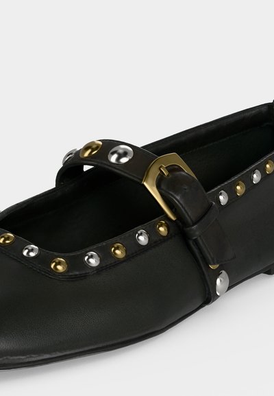 Chaussure plate noire avec une bride, ornée de clous ronds dorés et argentés et d'une boucle dorée, sur un fond clair et uni.