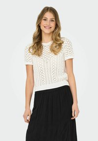 Top de punto blanco de manga corta con un patrón abierto, cuello redondo y dobladillo acanalado, combinado con una falda negra de volantes con detalles bordados.
