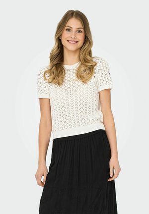 Top de punto blanco de manga corta con un patrón abierto, cuello redondo y dobladillo acanalado, combinado con una falda negra de volantes con detalles bordados.