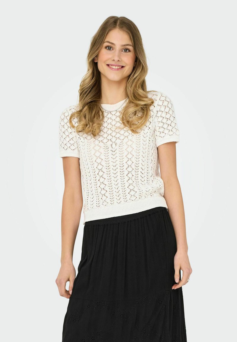 Top de punto blanco de manga corta con un patrón abierto, cuello redondo y dobladillo acanalado, combinado con una falda negra de volantes con detalles bordados.