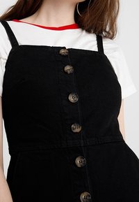 Svart denim ärmlös klänning med rak halsringning, med sex bruna knappar framtill och band. En vit skjorta med röd trim under.