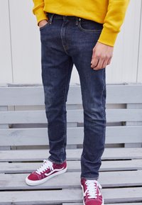 Jeans de mezclilla azul oscuro con un corte ajustado, con costuras naranjas, trabillas para cinturón y bolsillos delanteros. Combinados con zapatillas burdeos.