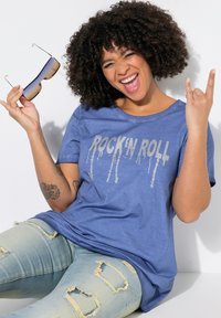 Modrá tričko s stříbrným designem textu "ROCK'IN ROLL", spárované s opotřebovanými světle modrými džíny. Model drží sluneční brýle a vyjadřuje hravou gestikulaci.