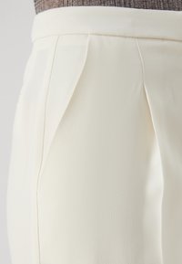 Pantalons plissés crème avec une texture lisse et une taille structurée, présentant un drapé subtil et un tissu léger.