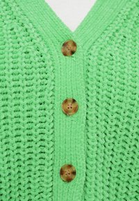 Πράσινη πλεκτή ζακέτα με λαιμόκοψη σε σχήμα V, που διαθέτει τέσσερα καφέ κουμπιά και υφή με μοτίβο ραφής. Μαλακό, βελούδινο ύφασμα.