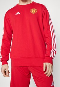 Uomo che indossa una felpa rossa del Manchester United e pantaloni con loghi e strisce bianche di Adidas sulle maniche e sui pantaloni.