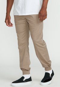 Khaki joggerki z elastycznymi mankietami, gładką fakturą i klasycznym krojem, zestawione z czarnymi butami typu slip-on i białymi skarpetkami.