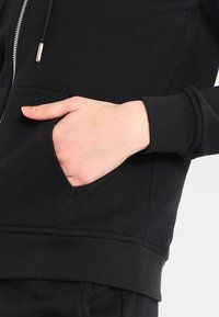 Crna zip-up dukserica s prednjim džepom, rebrastim manžetama i porubom, s glatkom teksturom tkanine i kontrastnim srebrnim detaljima na zipu.