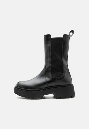 Bota de cuero negro hasta el tobillo con suela gruesa, panel elástico lateral y lengüeta para tirar, sobre fondo blanco.
