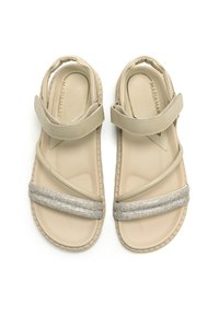 Sandalias beige con un upper suave y una correa plateada brillante. Presenta un diseño cruzado y una suela texturizada para mayor agarre.
