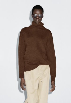 Bruine coltrui met een getextureerd breipatroon. Gepaarde met beige broeken. Het model draagt donkere zonnebrillen. Eenvoudige, minimalistische stijl.