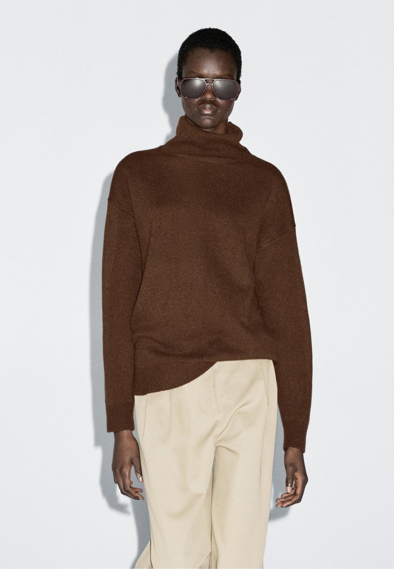 Bruine coltrui met een getextureerd breipatroon. Gepaarde met beige broeken. Het model draagt donkere zonnebrillen. Eenvoudige, minimalistische stijl.