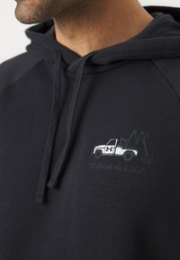 Hoodie noir avec une poche avant, comportant un graphique de camion blanc et le texte "Nous soulevons tous, Grands et Petits" en broderie grise.