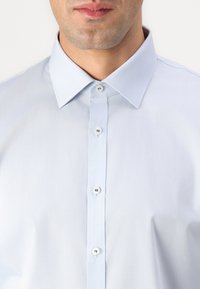 Cămașă albastră deschis cu guler clasic, având o textură netedă și cinci butoane frontale pentru închidere.