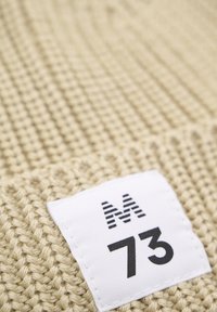 Beige színű kötött anyag, texturált felülettel. Egy fehér címke látható rajta, amelyen feketével az "M73" felirat olvasható, a jobb alsó sarokban elhelyezve.