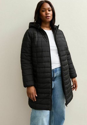 Zwarte pufferjas met een capuchon, voorzien van een gewatteerd ontwerp, ritssluiting en zijzakken, gedragen over een wit shirt en lichtblauwe jeans.