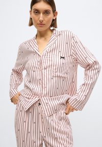 Set de pijamale din țesătură roz pal, cu dungi verticale subțiri. Include o cămașă cu nasturi și buzunar la piept, accentuată cu o panglică neagră.