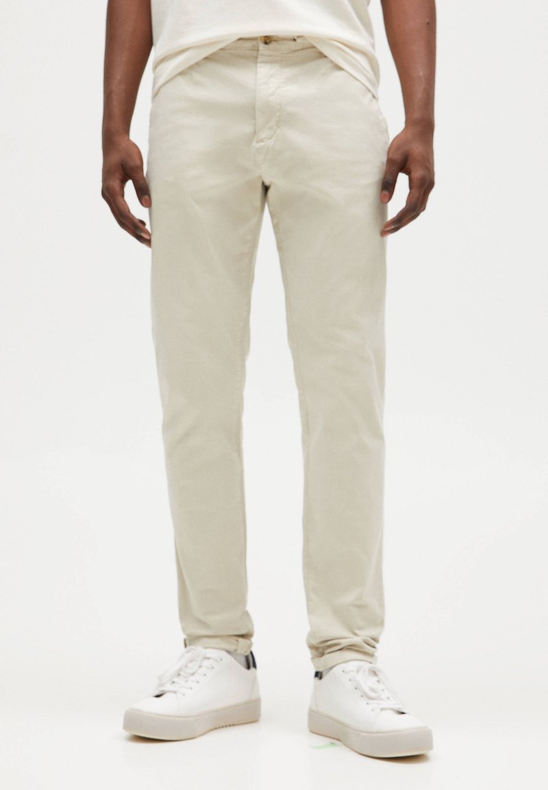Uomo che indossa pantaloni beige slim-fit e sneakers bianche, in piedi contro uno sfondo bianco uniforme.