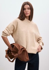 Bežna čez velikosti pulover s V-izrezom, zavihanimi rokavi in mehko teksturo; držan z rjavim suede usnjenim torbico; oblečen z temnimi kavbojskimi hlačami.