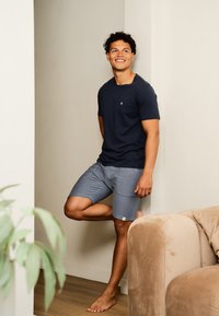 Navy t-shirt met een klein embleem, gecombineerd met lichtgrijze geblokte shorts. Model staat blootsvoets tegen een muur, met een pluche bank in de buurt.
