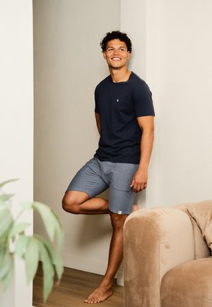 Navy t-shirt met een klein embleem, gecombineerd met lichtgrijze geblokte shorts. Model staat blootsvoets tegen een muur, met een pluche bank in de buurt.